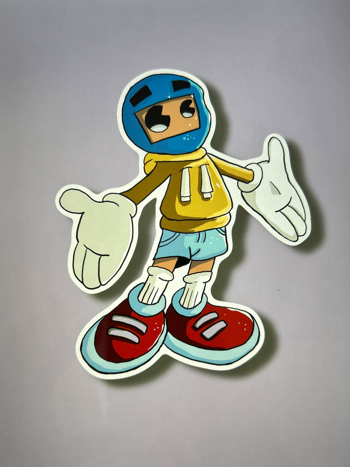A cartoon-style die-cut sticker of a character wearing a blue helmet, yellow hoodie, light blue shorts, and red sneakers, shown on a tablet screen.
